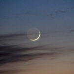 Young crescent moon an hour after sunset glowing through some clouds.
