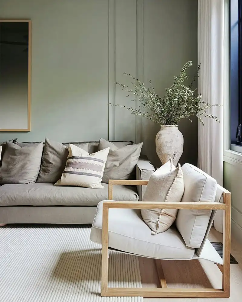 A neutral-toned living room with a light green paneled accent wall. In the foreground is a light wood armchair with white cushions, positioned on a textured white rug. Behind it sits a large, plush grey sofa with multiple taupe and striped cushions. A large, distressed ceramic vase holds green branches in the corner.
