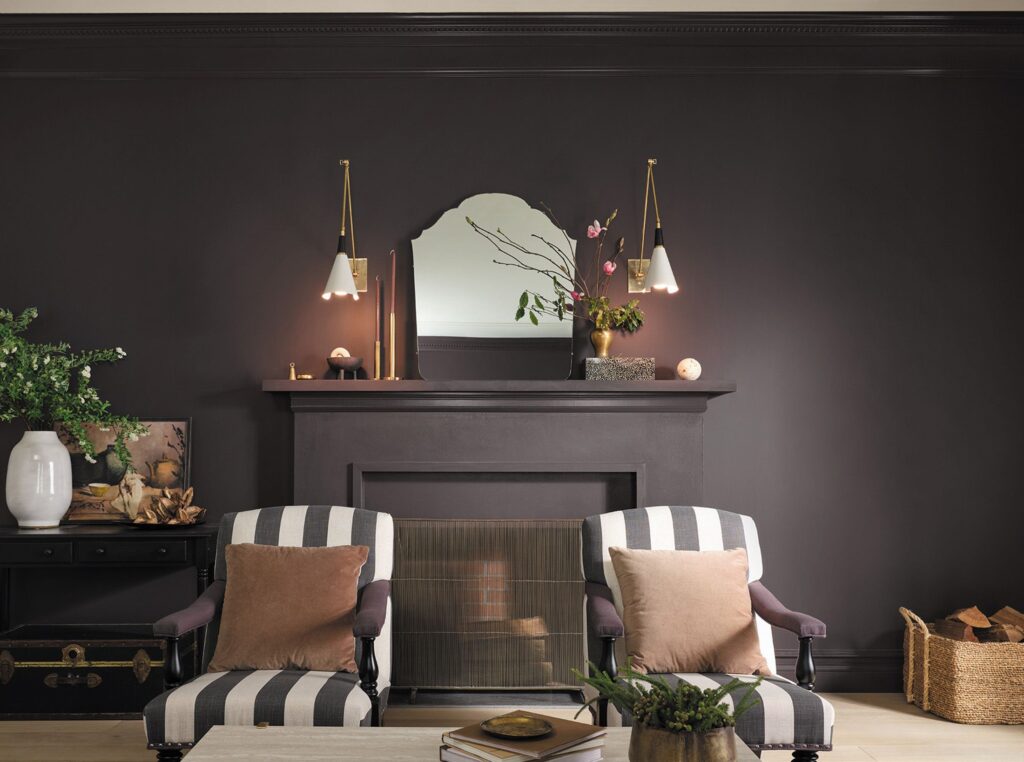 A dark and moody living room featuring a fireplace mantel painted in a deep, rich purple-black hue, identified as Sherwin-Williams Darkroom. Two striped armchairs with brown accent pillows sit facing a coffee table in front of the fireplace. Above the mantel, an ornate arched mirror reflects the room, flanked by two mid-century modern brass and white wall sconces.