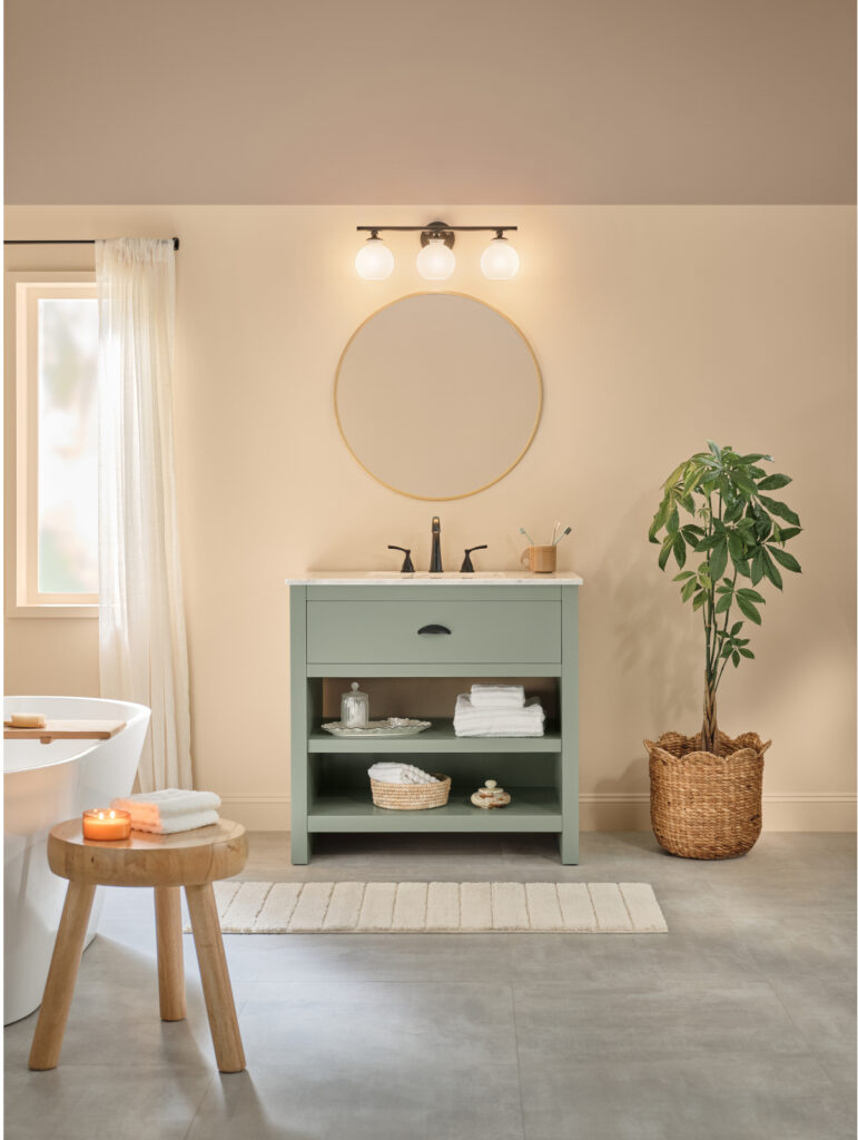 This image features a modern, transitional style bathroom with an earth toned colored walls with a sage green vanity, a round gold-framed mirror, and natural wood accents.