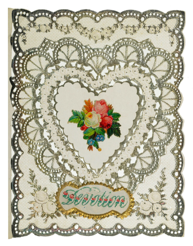A vintage Victorian Valentine's Day card, likely from the late 19th or early 20th century, featuring intricate multi-layered paper lace with silver and white detailing. The center of the card has a heart-shaped cutout revealing a colorful bouquet of roses and other flowers. Below the bouquet is an embossed gold frame with the word "Devotion" embroidered or printed inside. 
