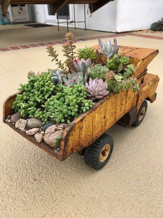 A rusty, vintage yellow Tonka dump truck repurposed as a creative garden planter. The truck bed is filled with a variety of lush green and pink succulents, including trailing varieties like burro's tail, and decorative river rocks at the back. The weathered, textured metal of the truck sits on a light-colored outdoor patio