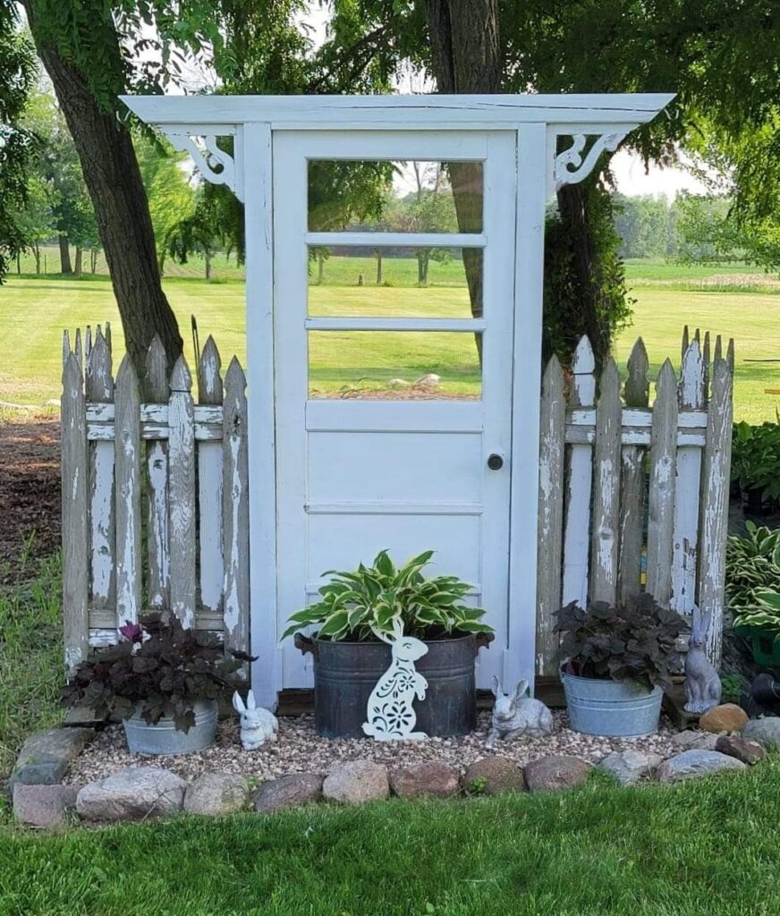 A charming garden decoration made of a white vintage door with glass window panes, flanked by short sections of weathered picket fence. The structure sits on a gravel base bordered by large stones, with potted plants and decorative rabbit figurines placed in front.