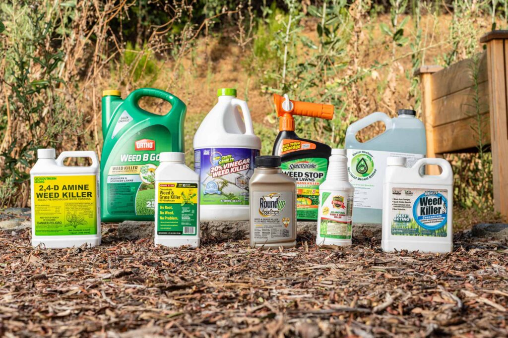 An outdoor, eye-level shot captures a collection of approximately nine weed killer containers arranged on a bed of brown wood mulch, with a blurred green hillside in the background. The containers vary in size, color, and brand, featuring names like Southern Ag, Ortho, Roundup, Spectracide, and Avenger. To the right, a portion of a wooden garden bed is visible. The scene is brightly lit by natural sunlight, casting soft shadows on the mulch.