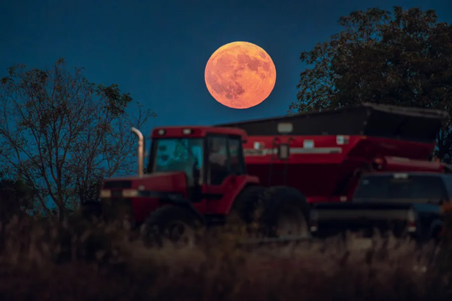 Red tractor in a field beneath a huge orange full moon, framed by trees at dusk
